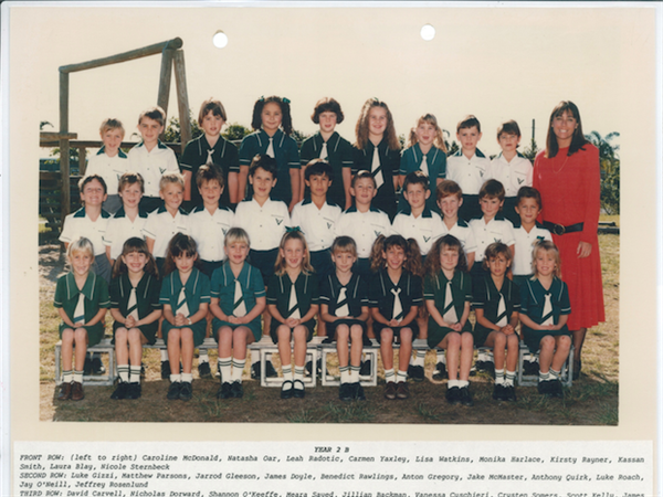 Historical class photo of primary school students in uniform with a teacher standing to the side, reflecting school community and past student life