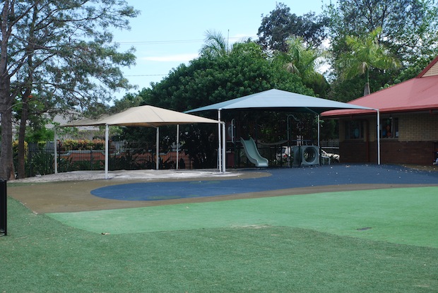 Shaded outdoor play area with soft flooring, play equipment and covered structures in a school setting
