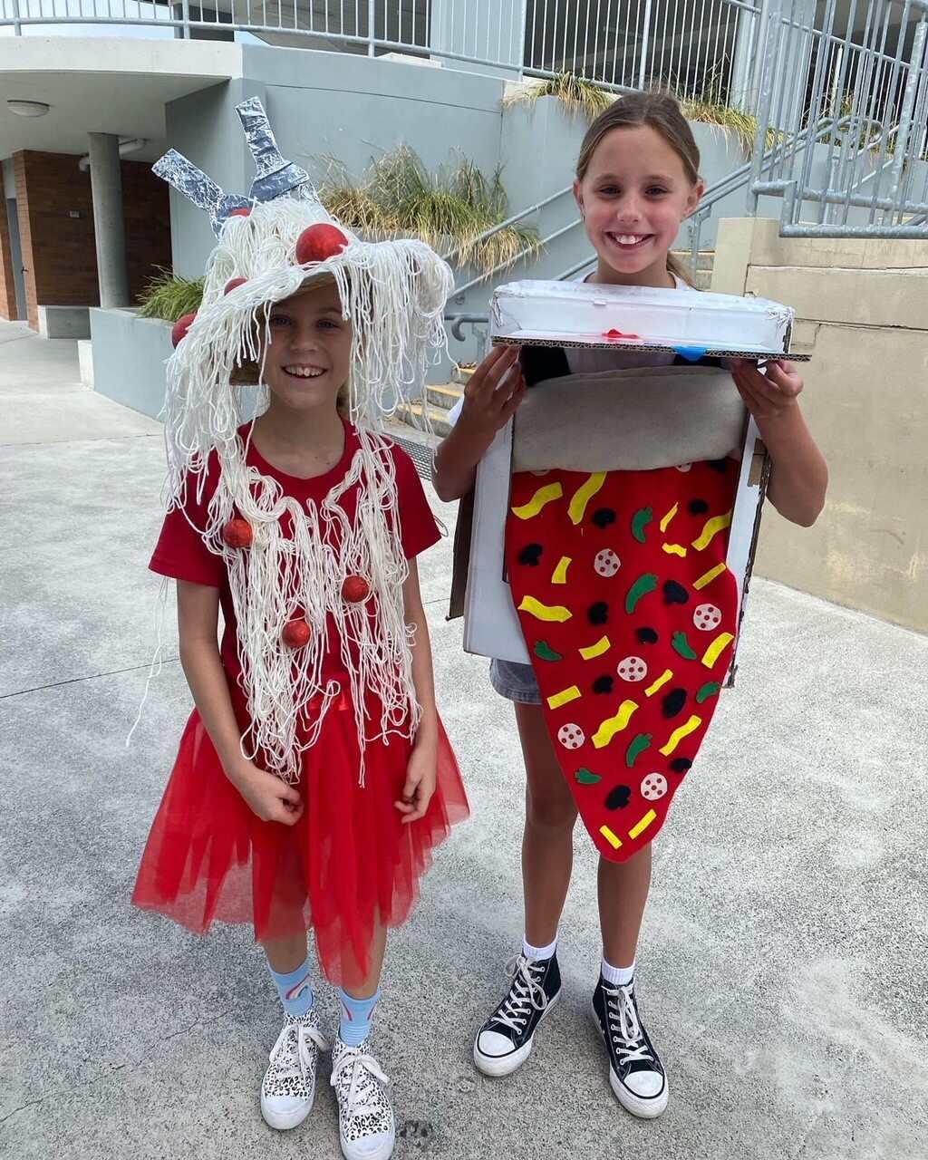Two students stand outdoors in costume, one dressed as spaghetti with a yarn headpiece and the other holding a cardboard pizza prop during a dress-up event
