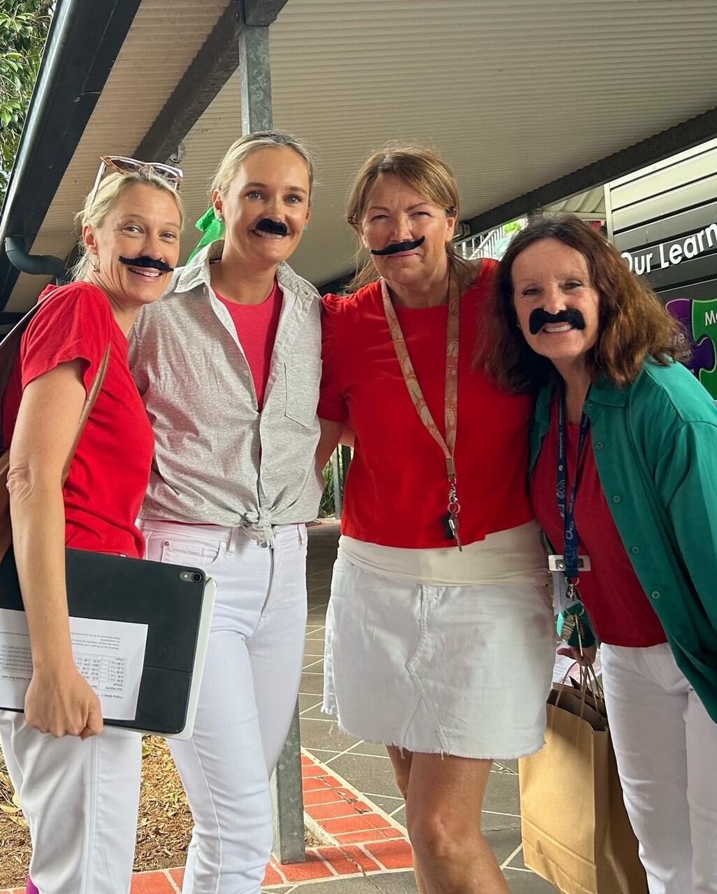 Staff members standing together outdoors wearing themed costumes with novelty moustaches during a school event.