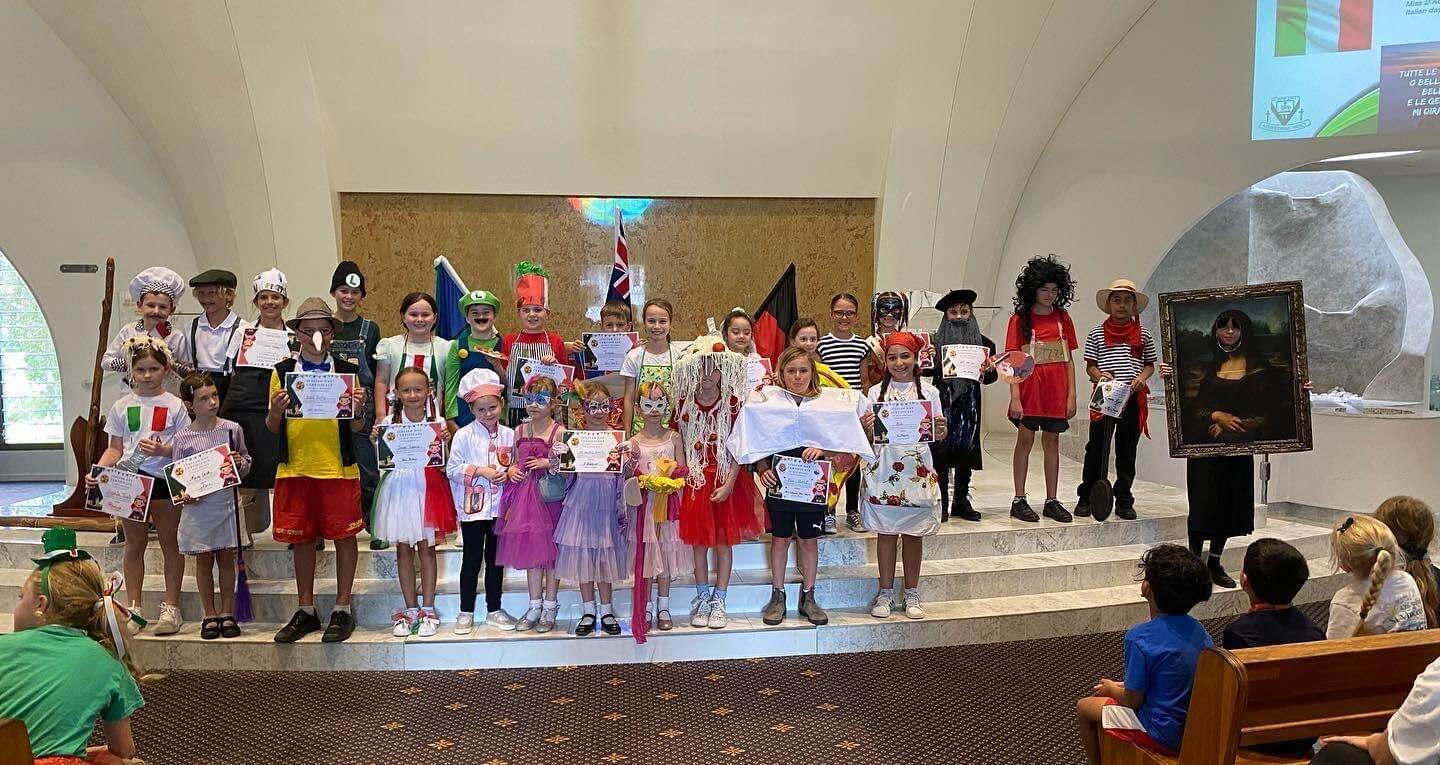 Primary school students in costumes on a stage holding certificates during a cultural or dress up event, with audience seated in front and flags displayed behind them