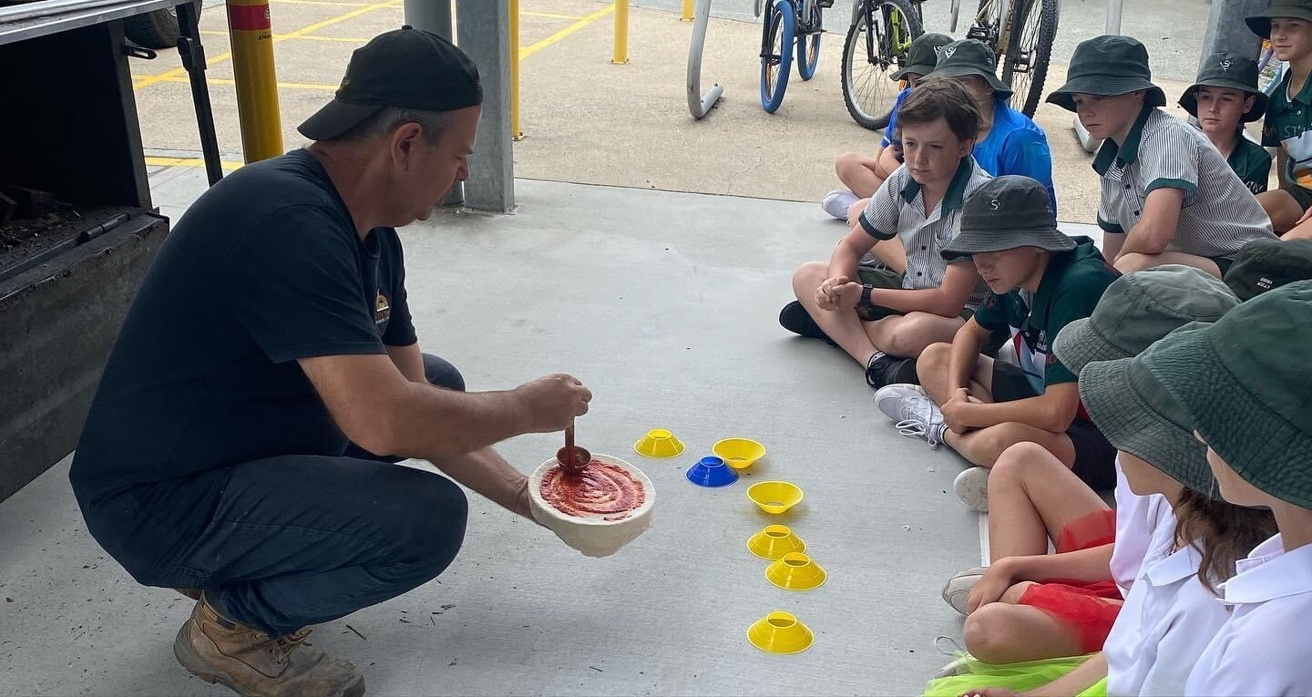 Students sit on the ground watching an adult demonstrate a hands-on activity using a bowl and red liquid, with small coloured cones placed in front during an outdoor learning session
