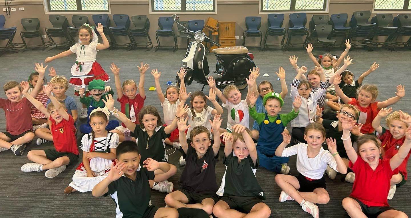 Students participating in a group activity, sitting together indoors with hands raised while a scooter is displayed at the centre of the space