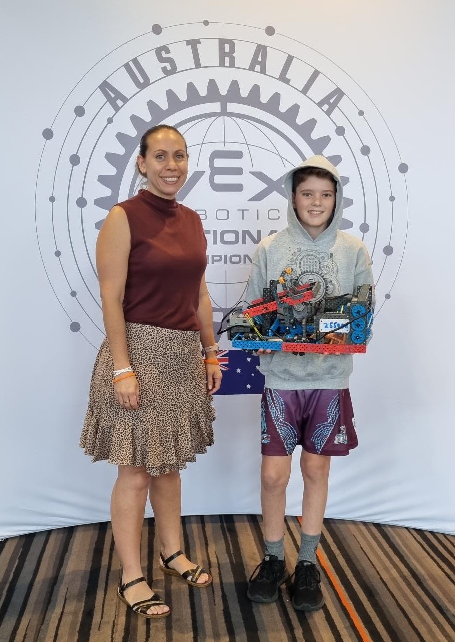 Student and staff member standing together holding a robotics project in front of a VEX Robotics competition backdrop