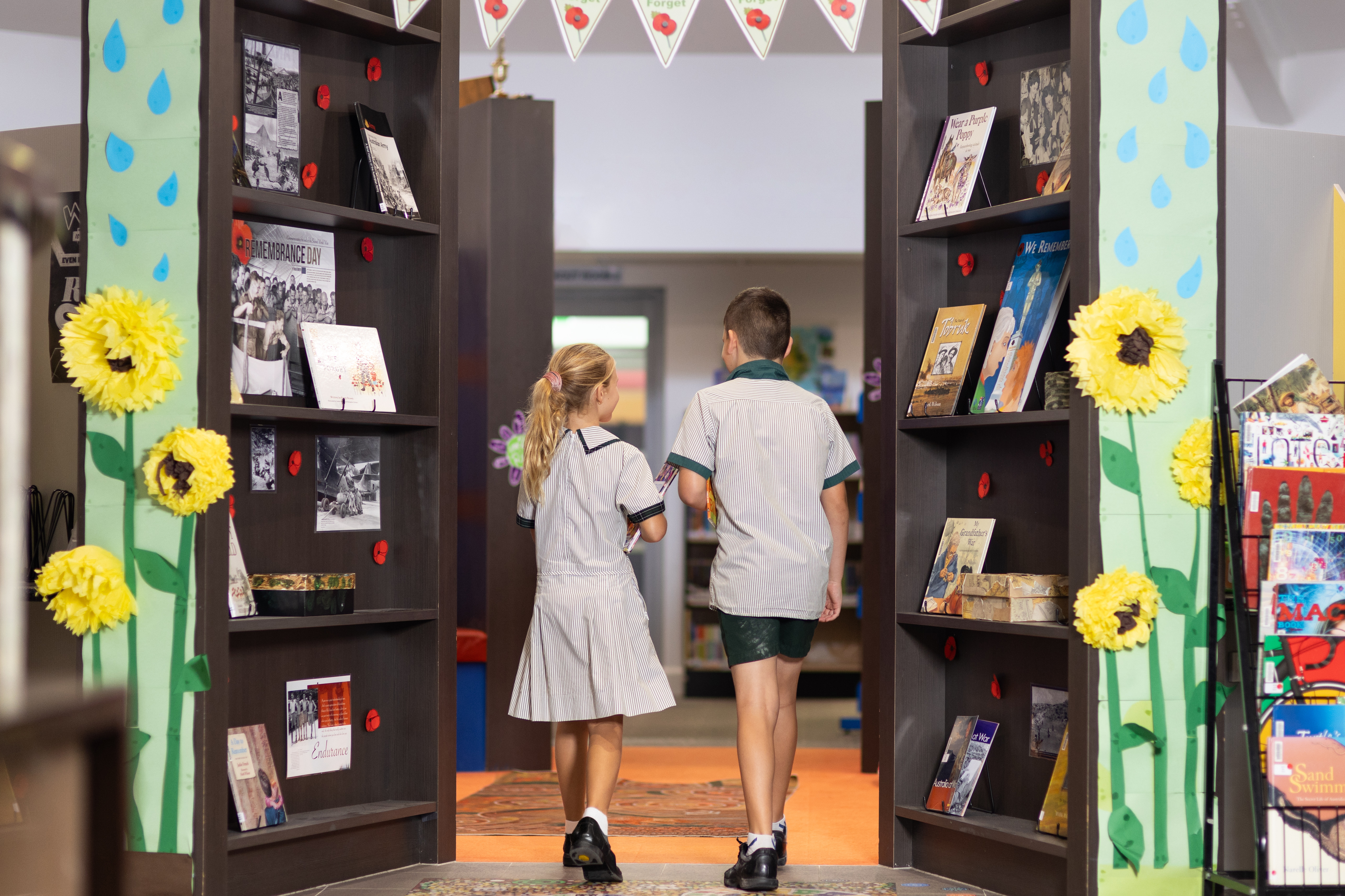Two students walking through a school library space between bookshelves decorated with learning displays and Remembrance Day materials