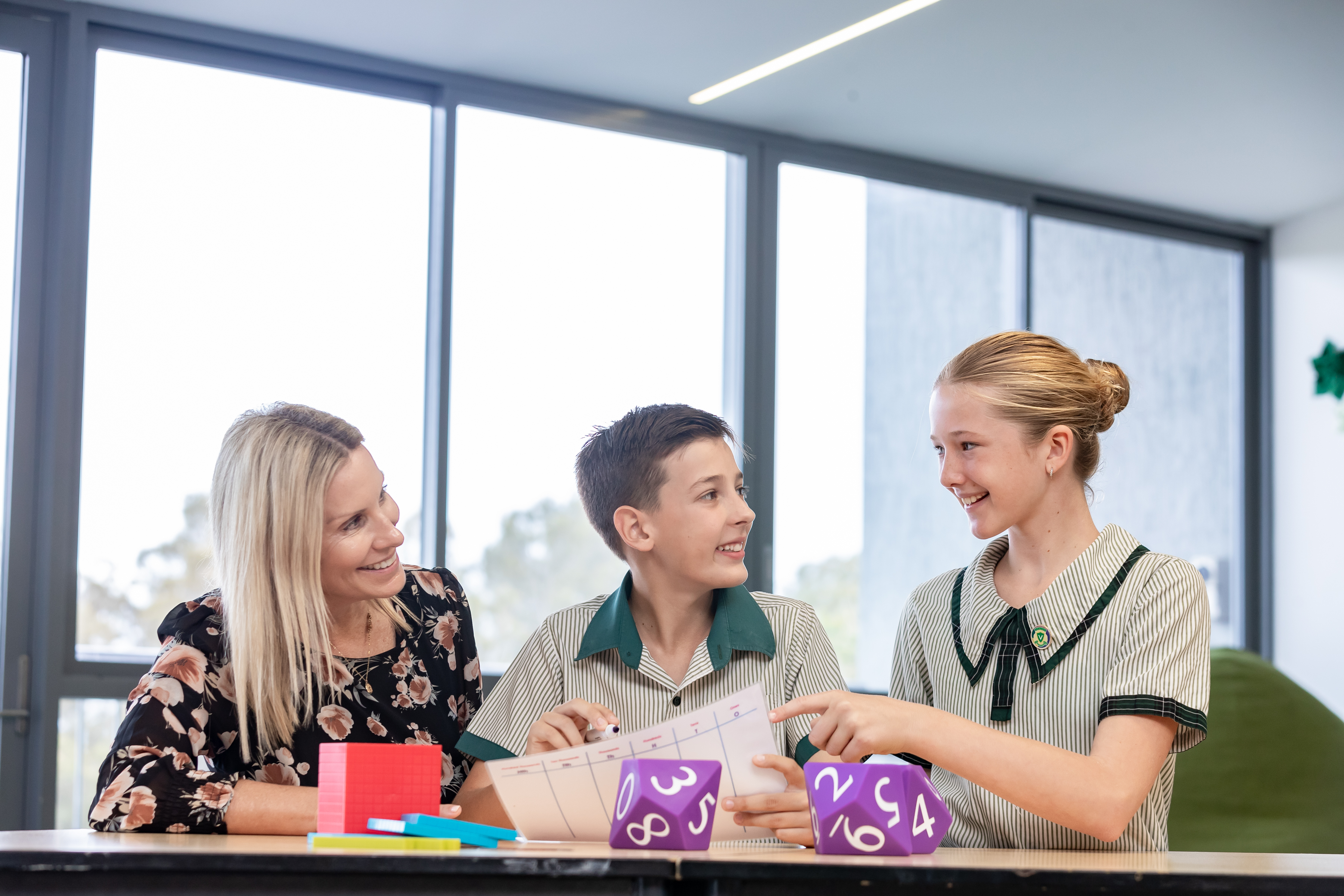 Teacher working with two primary school students at a classroom table using maths cubes and number chart while collaborating on an activity.