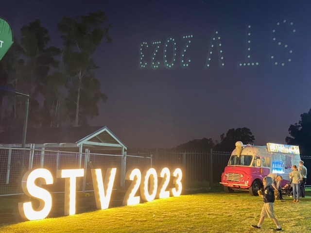 School community event at night with illuminated STV 2023 letters, families gathered on the grass, and lights forming a message in the sky